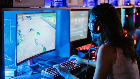 A young woman smiles as she plays video games in a computer room, wearing a headset.
