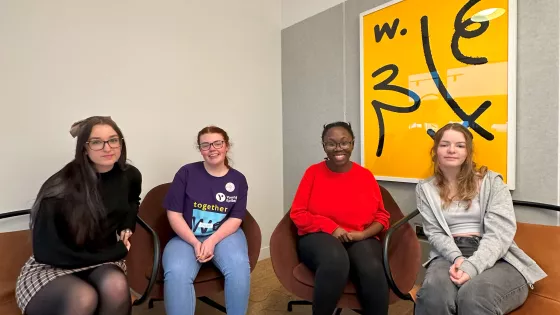 Four young women, one in Young Epilepsy branded clothes, sit before a large piece of art smiling.