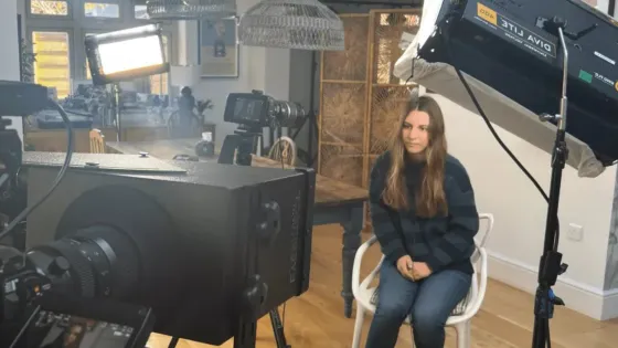 A woman is seated in a house facing a large camera.