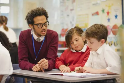 A teacher sits beside two primary school pupils working at a desk.