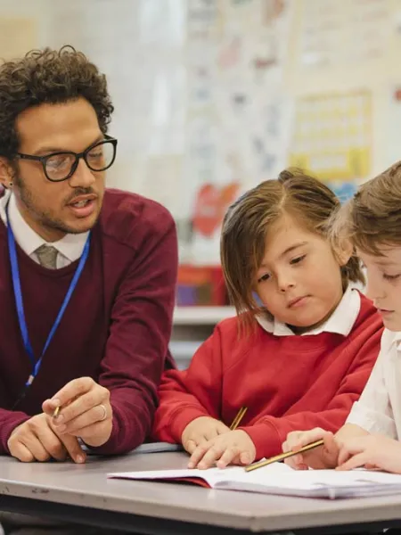 A teacher sits beside two primary school pupils working at a desk.
